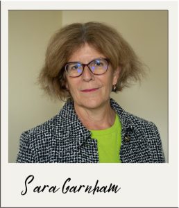 A polaroid frame containing picture of Sara Garnham - white female, with short dark blonde hair and glasses. wearing a green top and grey jacket